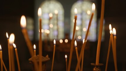 Trucking shot of thin lit candles in a European orthodox church with stained glass windows in the background