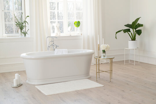 White modern bathroom with silver fittings with large sunny windows, decorations and plants. Interior design concept. Soft selective focus.