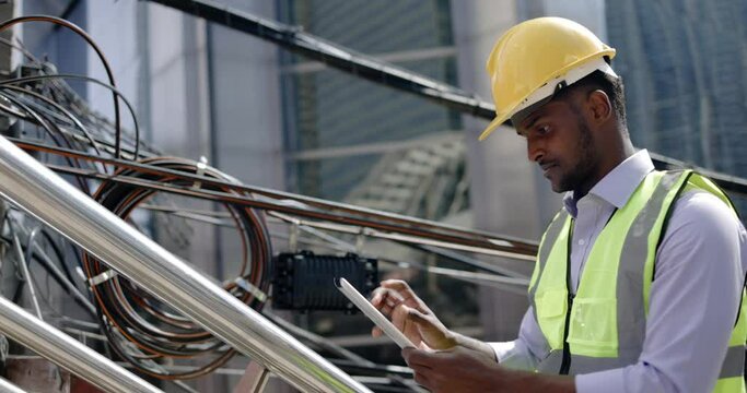 Electrical engineer checking electrical wires Internet cables on electric poles in city. African American technician check data. Repairing from tablet with expertise. supervise construction site