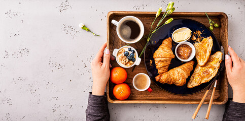 Woman serving continental breakfast on wooden tray. Coffee, croissants, jam, butter and fruits.