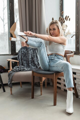 Stylish beautiful young blonde girl in fashionable clothes with jeans dresses a shoe and sits in an wooden vintage chair in a room