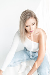 Obraz na płótnie Canvas Delicate beautiful young blonde woman with a white t-shirt and vintage blue jeans sits on a white background in sunlight