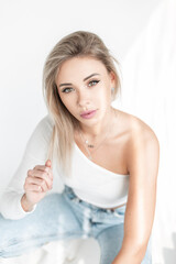 Obraz na płótnie Canvas Cute blonde woman in fashion white clothes with t-shirt and blue jeans sits in studio at sunlight