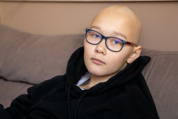 portrait depression sad bald young teen girl with cancer after chemotherapy. wearing glasses and black hoodie, sitting on the couch. - obrazy, fototapety, plakaty