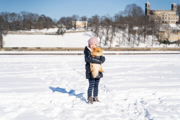 Fototapeta na wymiar Portrait of young little girl in warm jacket with fur, jeans, boots and gloves holding on her hands, hugging dog pet Pomeranian Spitz on sunny winter day, in snowy white field with blue sky. Holiday