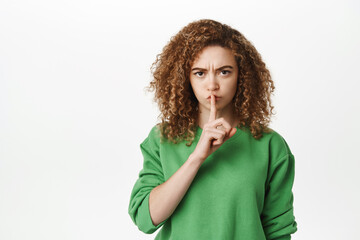 Portrait of caucasian girl with curly hair, showing shhh, shush hush gesture, press finger to lips, be quiet sign, standing over white background