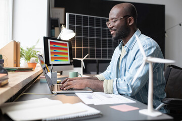 African man in eyeglasses sitting at his workplace with documents and typing on laptop during his work at office