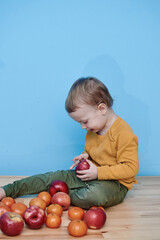 Fototapeta premium a little boy of 2 years old sits on a table against a blue wall and holds a red apple. there are a lot of red apples and a lot of tangerines around. child in green pants and yellow sweater