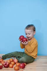 Fototapeta premium a little boy of 2 years old sits on a table against a blue wall and holds a red apple. there are a lot of red apples and a lot of tangerines around. child in green pants and yellow sweater