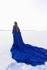 Incredible stunning girl in a blue airy dress with a long train. In the background winter landscape frozen river covered with ice. The woman in the dress is standing with her back