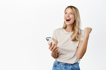 Enthusiastic young woman winning on mobile phone, rejoicing, using smartphone app, celebrating, triumphing on cellphone, white background - 485865241