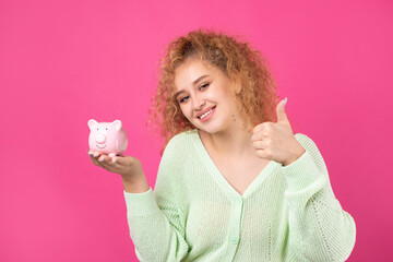 A cute young girl with curly red hair holds a piggy bank, a pink piglet in her hands. The concept of wealth and safety of money.