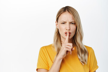 Naklejka premium Portrait of angry blond girl shushing, hush with upset frowning face, making be quiet, silence gesture, standing in tshirt over white background