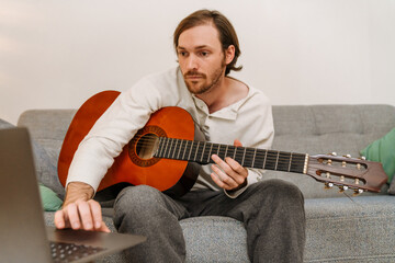 Ginger man playing guitar and using laptop while sitting on couch