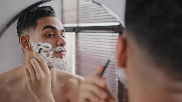 Hispanic arabian arab indian bearded handsome man shaving using disposable razor in bathroom looking at mirror. Daily morning routine male hygienic beauty procedure. Guy shaves beard and moustache