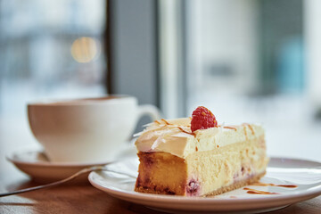 Cup of coffee and piece of raspberry cheesecake on cafe table near window, breakfast at public place