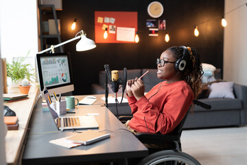African woman in headphones sitting at the table with laptop and speaking to the microphone during...