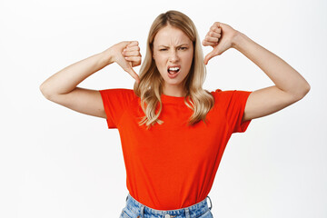 Disappointed blond girl shows thumbs down, dislike, disapproval sign, negative feedback, standing...