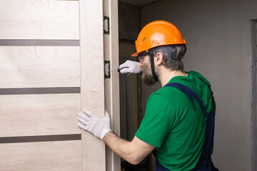 A male locksmith installs a door in an apartment. The guy works with a screwdriver. - obrazy, fototapety, plakaty