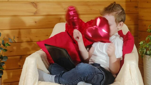 Valentine's Day children or kids. Cute little boy chatting via video call, on a laptop, gadget, on Valentine's Day, a red heart-shaped balloons in his hands. Congratulation or postcard for Valentine's