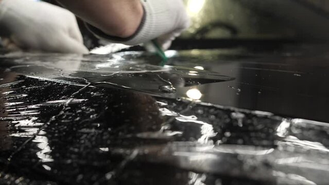 Worker sticking anti-gravel film on a black car body with scrapper at the detailing vehicle workshop, close-up. Concept of car body protection with special films.