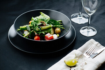 Tasty greens, vegetables and meat beautifully plated in a single dish. In a black bowl over black tablecloth. Cutlery at sides and couple of empty glasses.