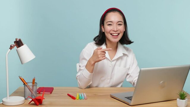 Young successful employee business woman of Asian ethnicity 20s in casual shirt sit work at office desk point on pc laptop show thumb up isolated on plain pastel light blue background studio portrait
