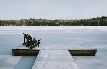 Snow covered dock with adirondack chairs on frozen lake in winter.