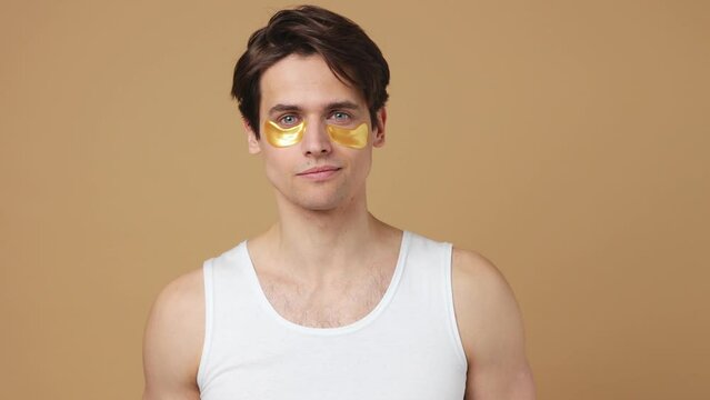 Satisfied young man 20s perfect skin wears white tank top put on wear gold patch under eye isolated on plain pastel beige background studio portrait. Body care healthcare cosmetic procedures concept