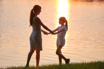 Fototapeta premium Happy mom and daughter girl relaxing holding hands enjoying time together in summer park at sunset. Family love and relationship concept
