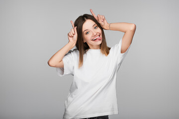 cheerful and tricky woman sticking out tongue and showing horns gesture isolated on grey