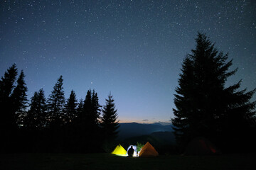 Silhouette of lonely hiker resting besides burning bonfire near illuminated tourist tents on camping site in dark mountains under night sky with stars. Active lifestyle and outdoor living concept