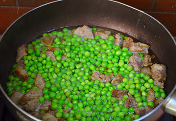 pork meat and a lot of young green peas are stewed in a large frying pan. steam is coming from the food being cooked