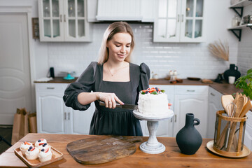 Pastry chef confectioner young caucasian woman in gray dress with knife cut slice cake on kitchen table. Cakes cupcakes and sweet dessert Scandinavian style studio