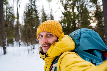 Fototapeta na wymiar Portrait of a broadly smiling traveler. Bright winter clothes and a hiking backpack behind him. Happy eyes and steam from the mouth. Hiking in the woods in winter