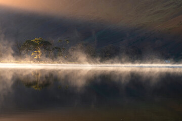 Misty sunrise with row of Pine trees giving perfect reflections in Buttermere, Lake District, UK. Beautiful rays of light shining on to side of mountain.