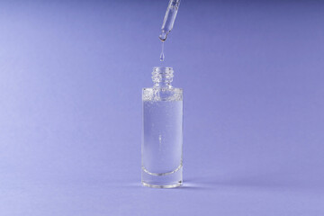 Bottle of cosmetic product and a glass pipette with a drop of cosmetic product on a purple trendy background.