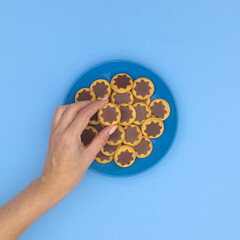 Woman hand taking one delicious chocolate and biscuit cookie from blue plate on cyan background. Food flat lay.