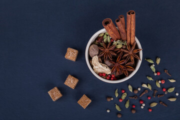 Spices cinnamon sticks, cardamom pods, star anise, sugar, nutmeg, coriander and pepper seeds in brown ceramic cup on a black slate stone plate. Spices for Masala tea.