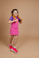 Active, excited young girl holding two kanekalon pigtails indulging trying to dance on one place with big smile with missing tooth in pink jumpsuit and purple t-shirt on beige background.
