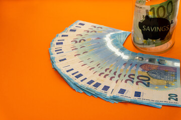 Blue banknotes of twenty euros stacked in fans on an orange background and a transparent glass piggy bank with one hundred euros inside and a pig in black printed on the glass with the word savings.