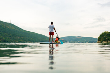 Back view of father with daughter in swim life vest swimming on SUP board. Family paddleboarding on...