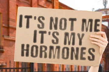 The phrase " It’s not me, it’s my hormones " on a banner in men's hand with blurred background. Sexy. Flame. Desire. Passion. Sex. Clinical. Action. Life. Energy. Pill. Test. Drug. Clinic. Growth
