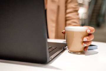 Close-up of beautiful female hands holding a large white cup of cappuccino. A business woman sits in a cafe working at a computer.