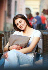 Portrait of joyful girl on street, sits dreamily in armchair on summer terrace of cafe, tourist city on background of crowd of people