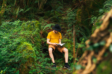 Naklejka na ściany i meble Caucasian man sitting in the middle of the jungle reading a book.