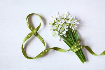 Geeting card for Women's day march 8, number eight from a green ribbon and a bouquet of snowdrops on a white wooden background - 485913025