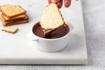 Crackers or cookies in chocolate paste in white sauce pan. A hand holds a cookie.