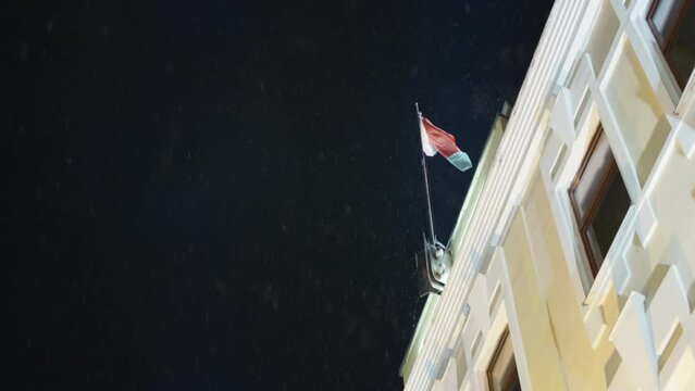 Bottom view of building with flag at night. Action. Building with flag on background of night sky. Beautiful snow falls at illuminated building at night