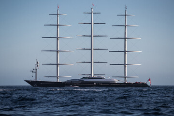 A huge sail yacht of conceptual design with three masts stands in the bay near Monaco at sunny day, the sails are lowered, the yacht spins around its anchor, left side of the sailboat, clear weather